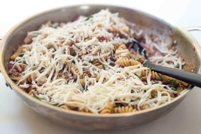 Shredded cheese on top of pasta and other ingredients in a skillet with a spoon.