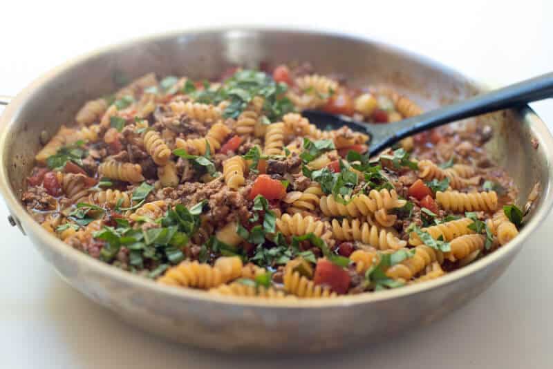 A spoon combining pasta with other ingredients in a skillet.