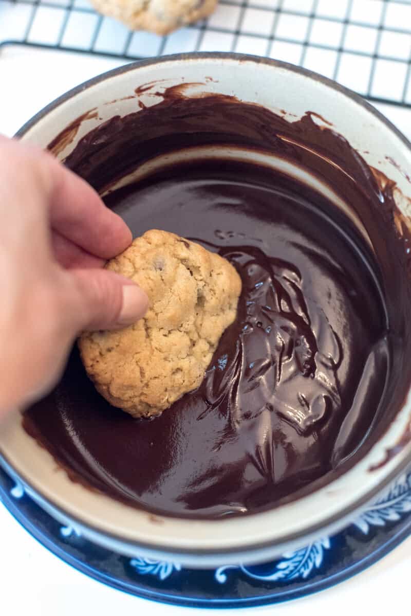 A hand dips a cookie into the melted chocolate mixture in a saucepan.