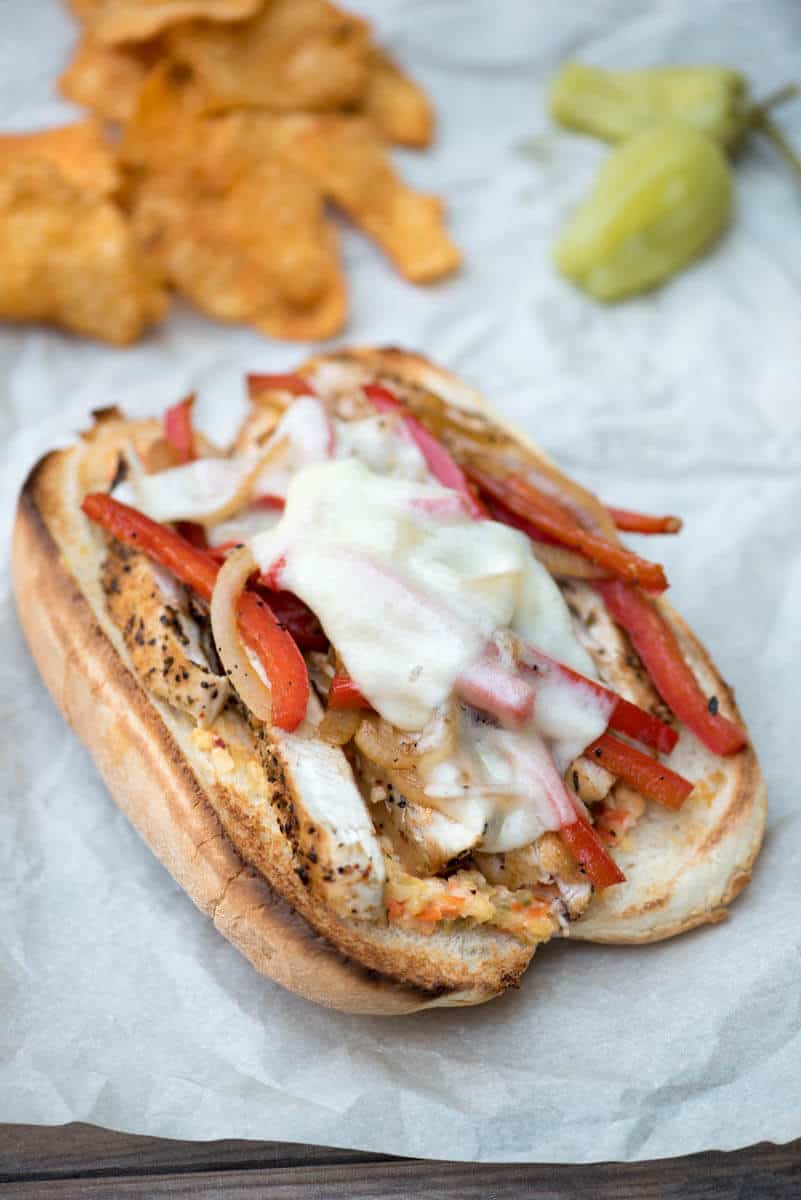 An Italian Chicken Sub on parchment paper with potato chips and green peppers in the background.