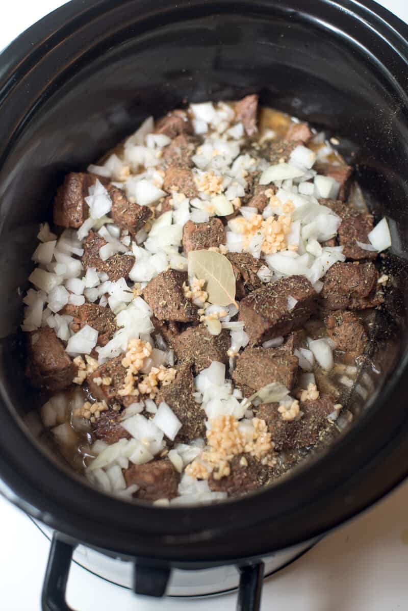 An in process shot of diced onion , garlic and a bay leaf over the beef tips in the slow cooker.