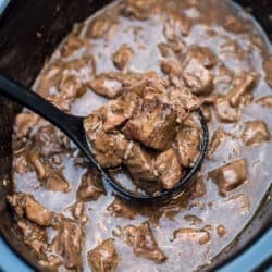A ladle scooping beef tips with gravy from a slow cooker.