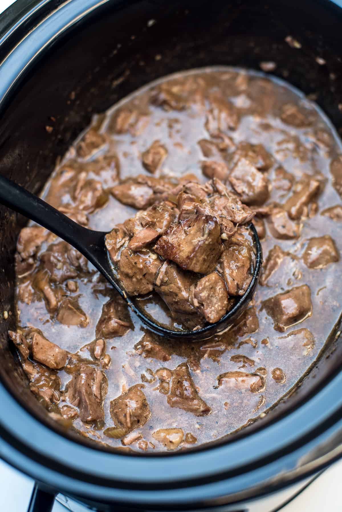 A ladle scooping beef tips with gravy from a slow cooker.