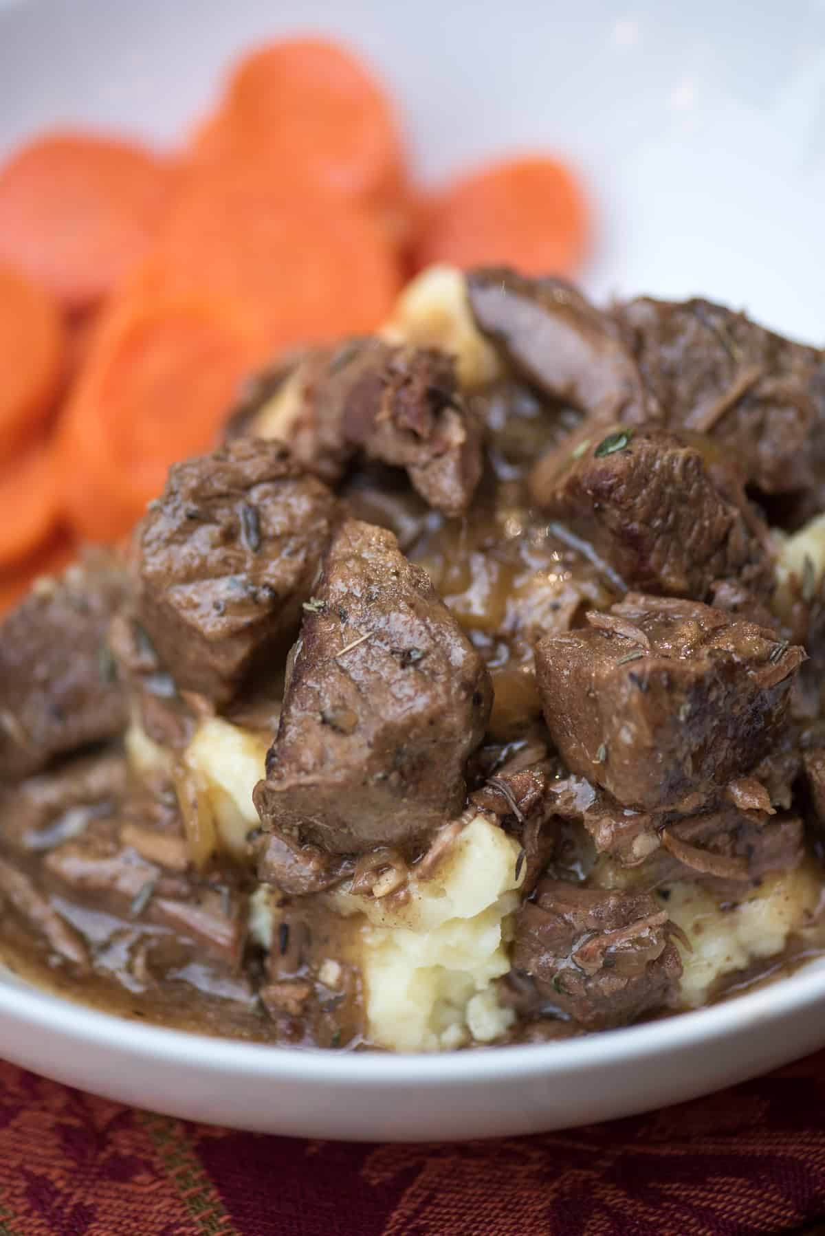 A close up of beef tips with gravy over mashed potatoes in a white bowl.