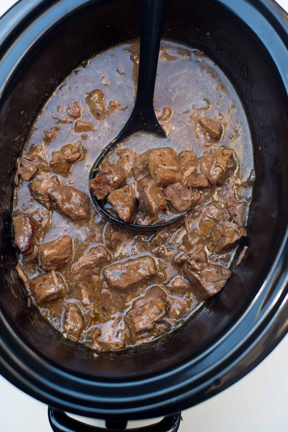 A ladle scooping beef tips with gravy from a slow cooker.