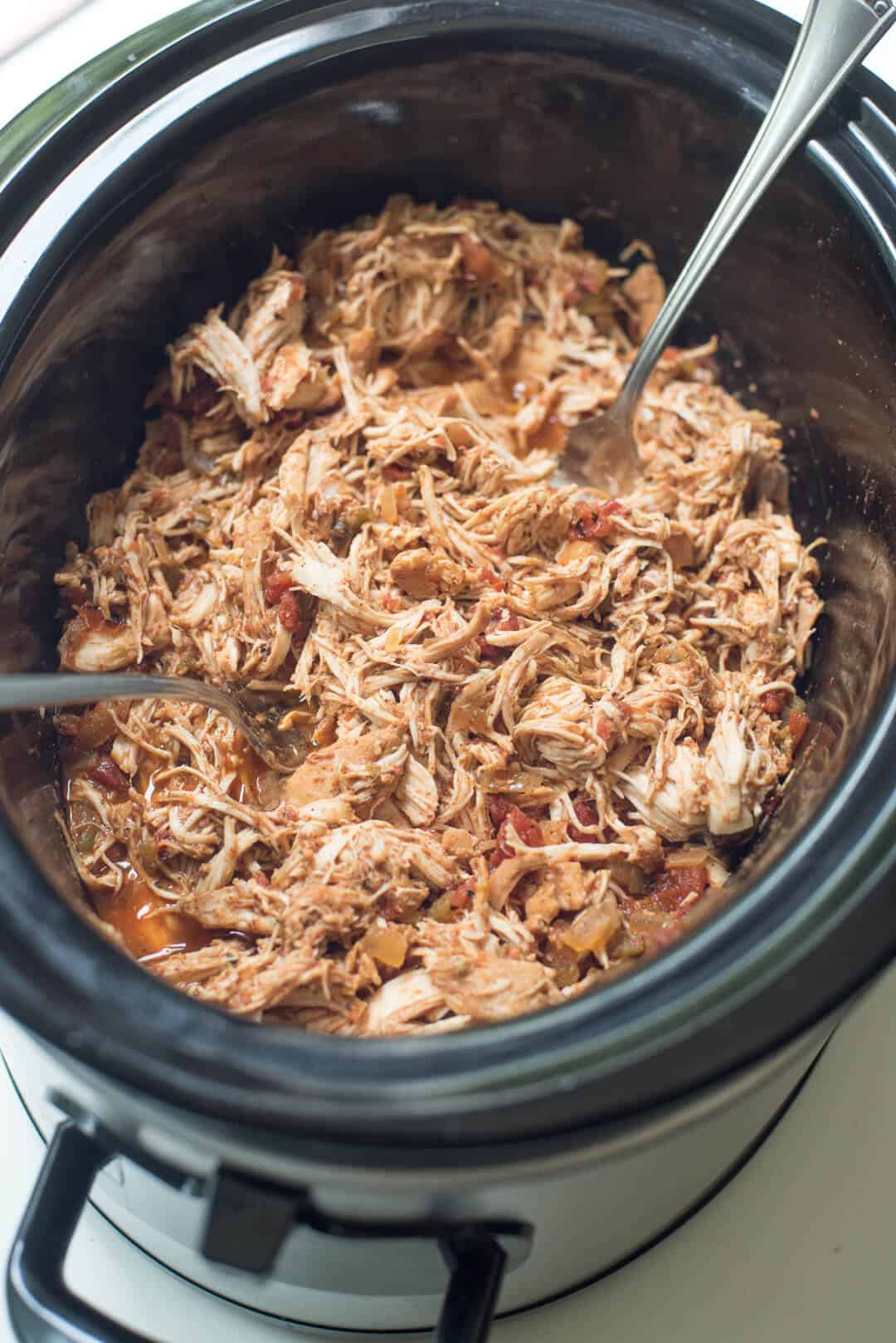 Cooked chicken being shredded with two forks in a slow cooker.