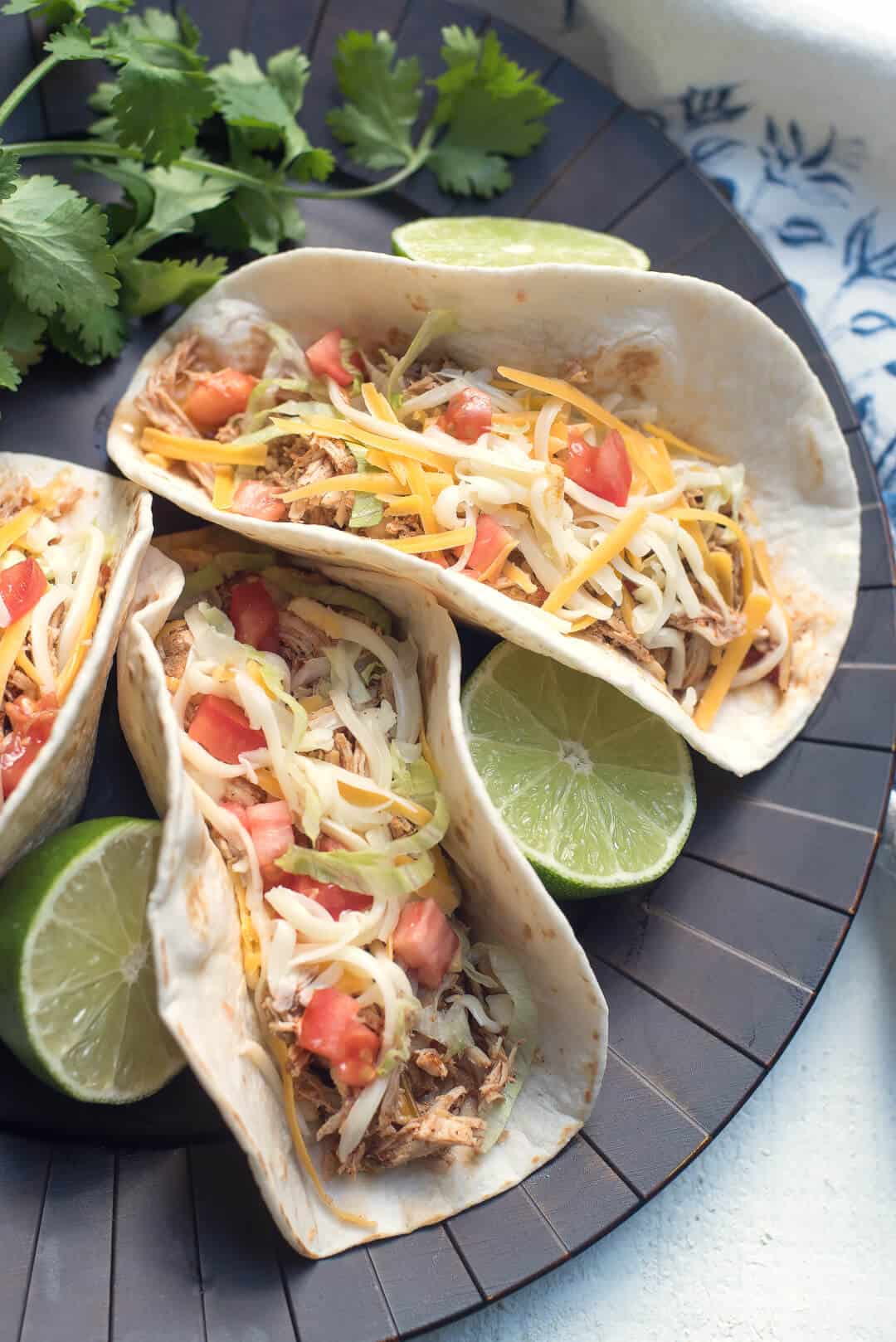 Three Crock Pot Chicken Tacos on a brown tray with limes and cilantro.