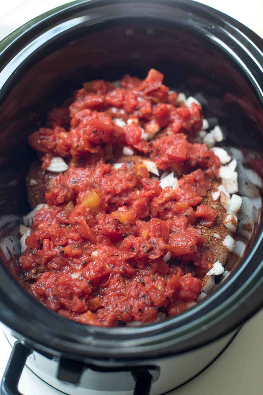Raw seasoned chicken in a slow cooker topped with diced onion and fire roasted diced tomatoes.