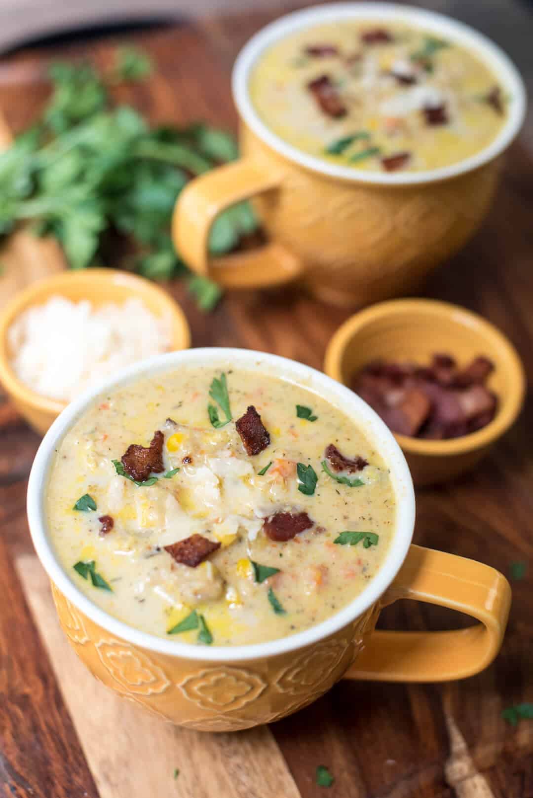 Mugs of Roasted Cauliflower Corn Chowder on a board with small bowls of Parmesan and bacon.
