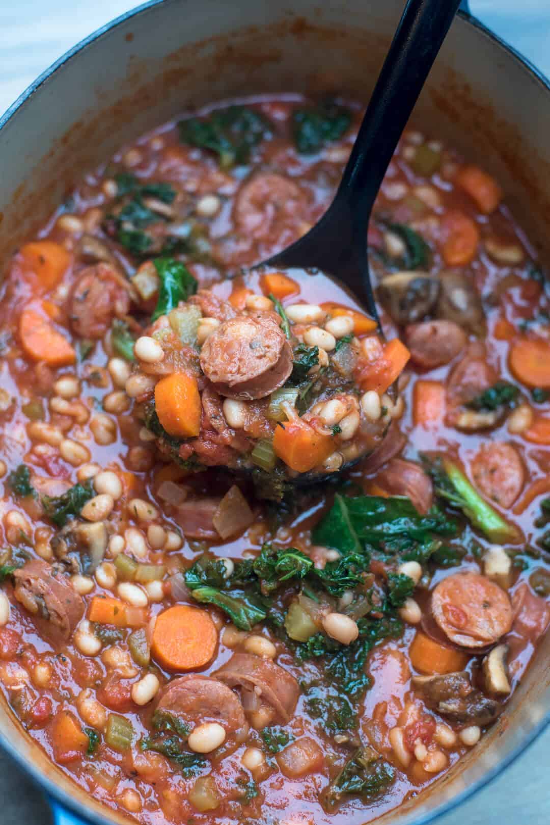 A ladle scoops Sausage White Bean and Kale Stew from a Dutch oven.