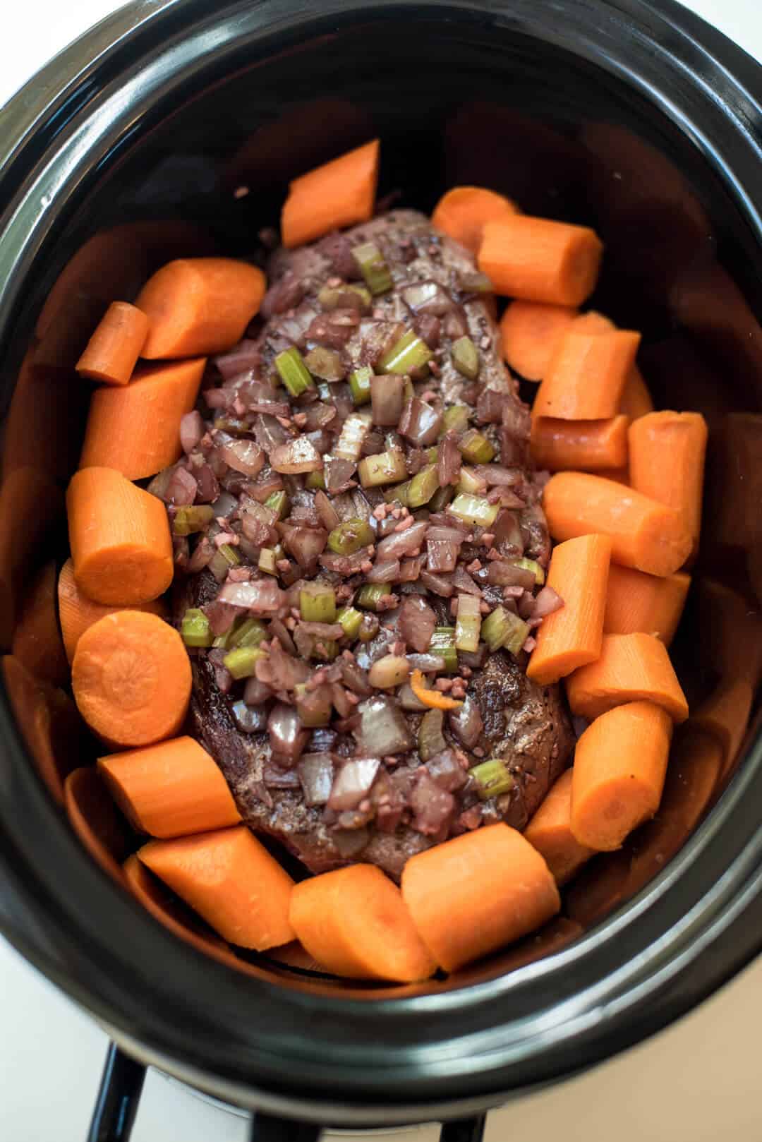A pot roast in a slow cooker topped with an onion and celery mixture and big chunks of carrot.