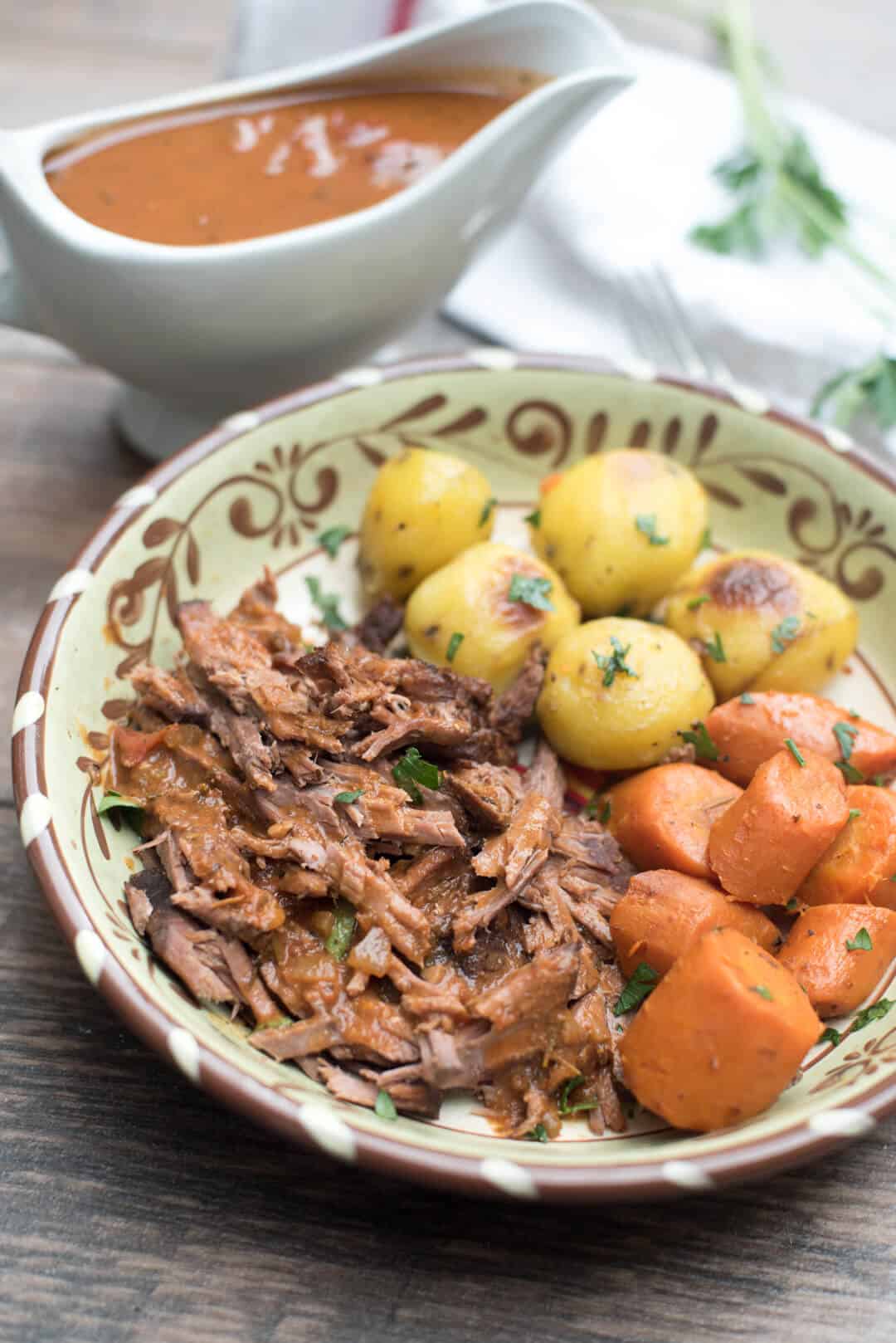 Italian Pot Roast in a bowl with carrots and potatoes in front of a gravy boat.