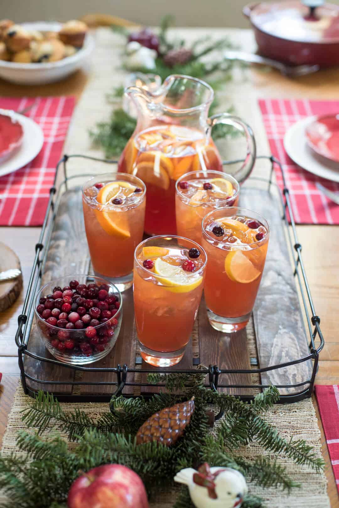 A wooden tray on a table with iced tea in glasses and a pitcher next to a bowl of cranberries.