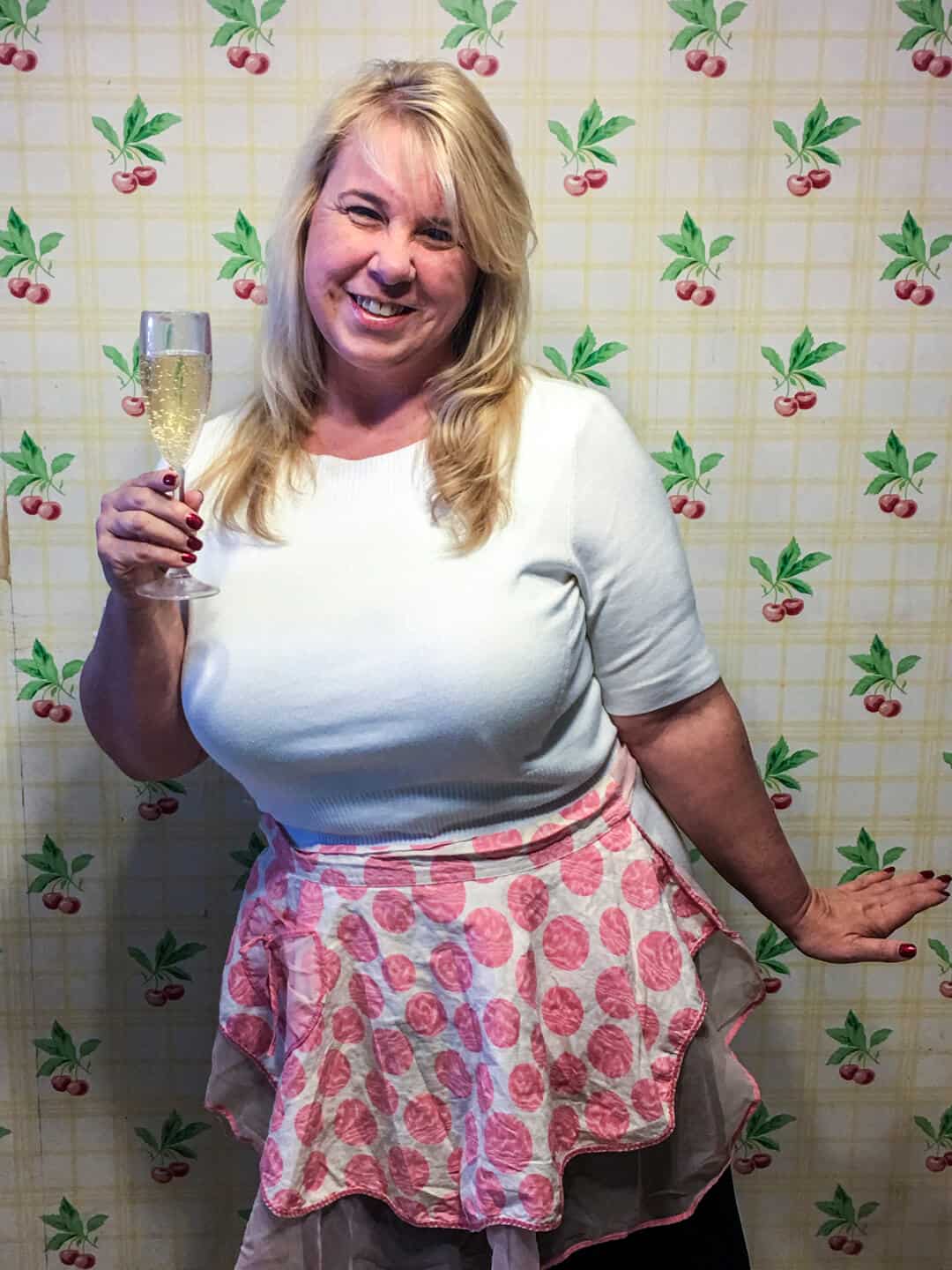 A blond woman wearing a pink apron and holding a glass of champagne.