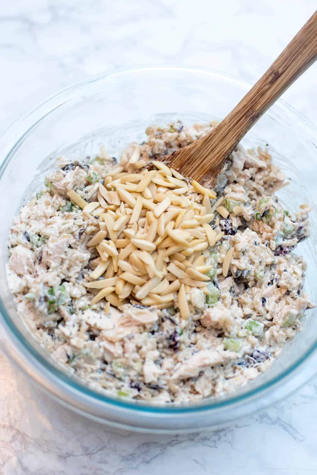 Slivered almonds being stirred into a chicken salad mixture in a glass bowl.