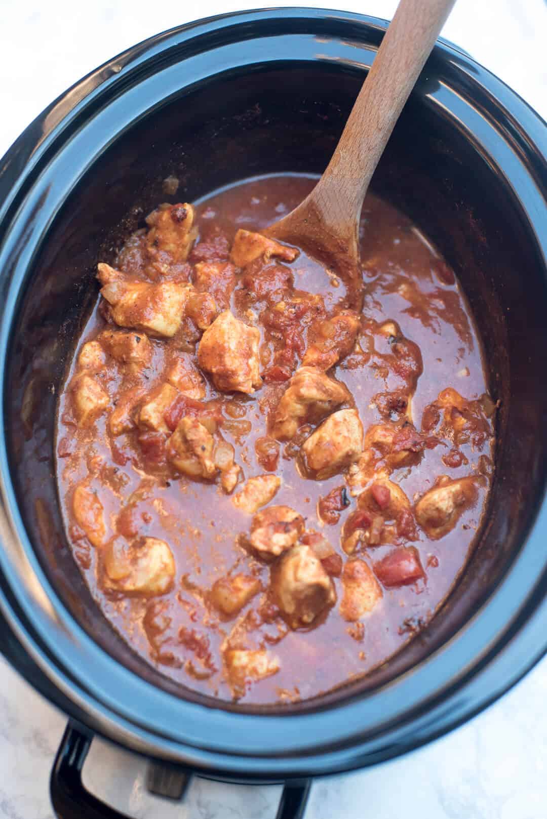 A spoon stirs cooked chicken and sauce in a slow cooker.