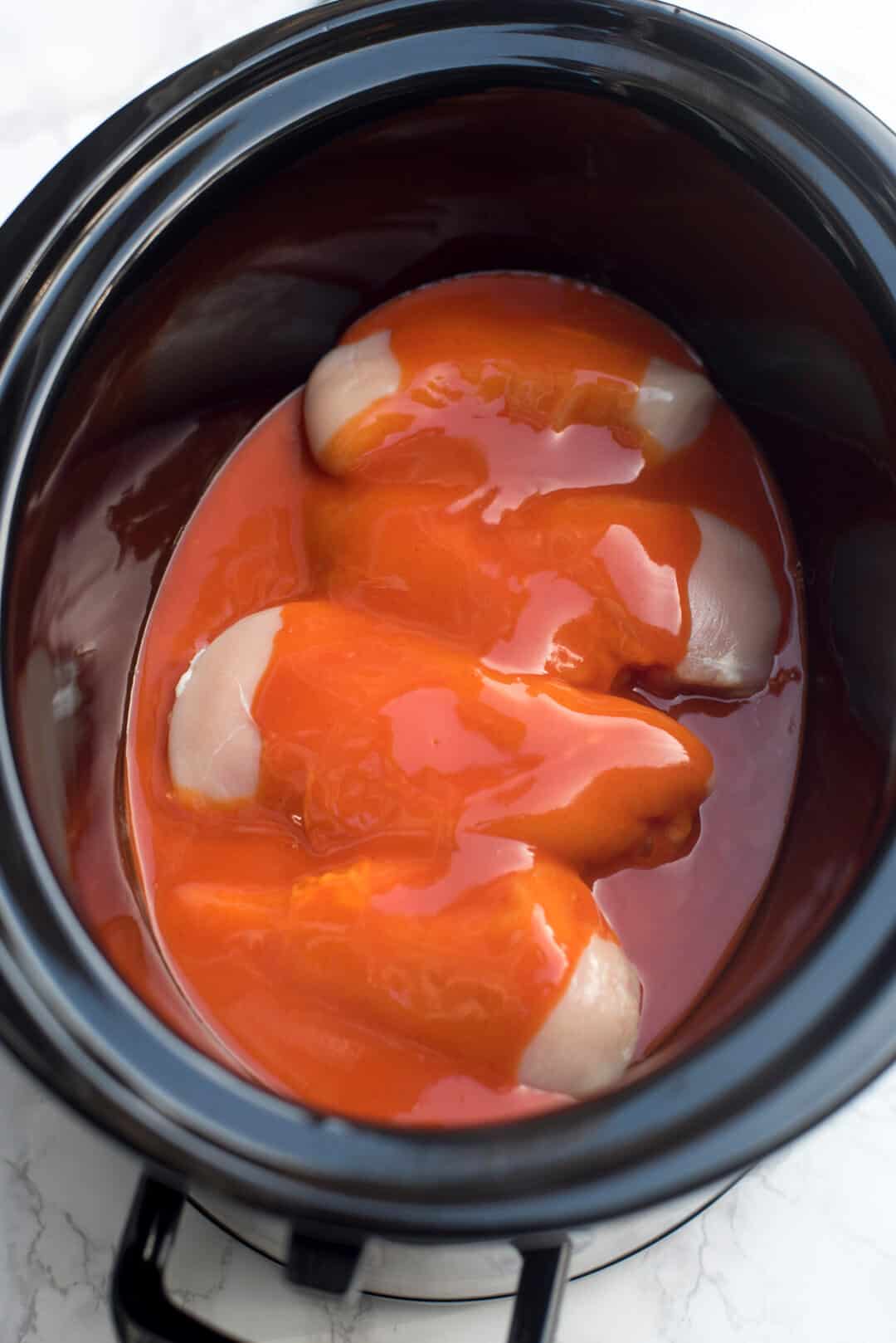 Raw chicken in a slow cooker topped with Franks Redhot Buffalo Wing Sauce.