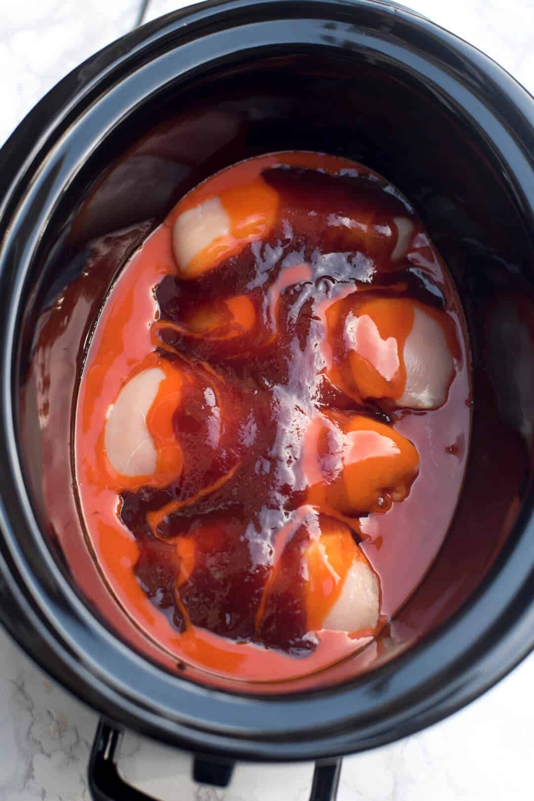 Raw chicken in a slow cooker topped with wing sauce and BBQ sauce.