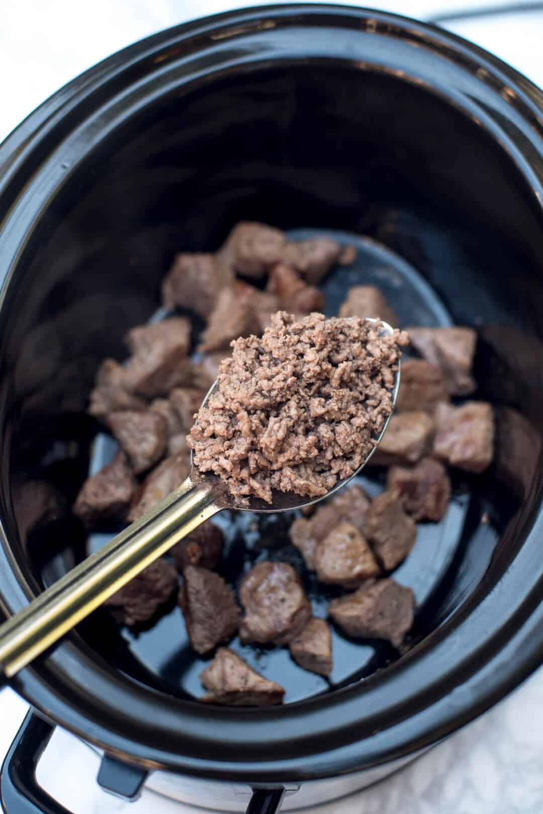 A large spoon holding cooked ground beef hovering over a slow cooker.