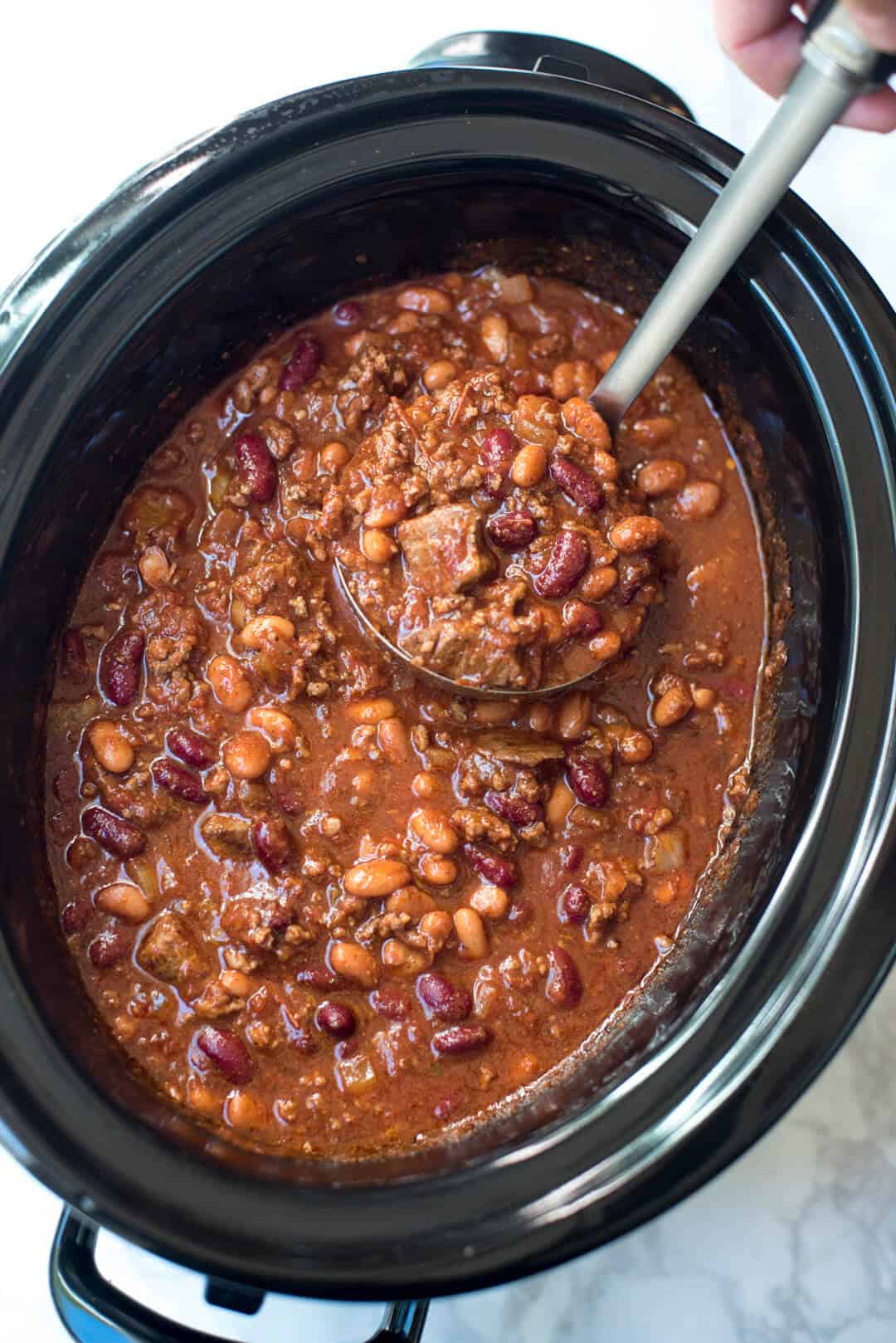 A ladle scoops chili with chunks of beef, ground beef and beans from a slow cooker.