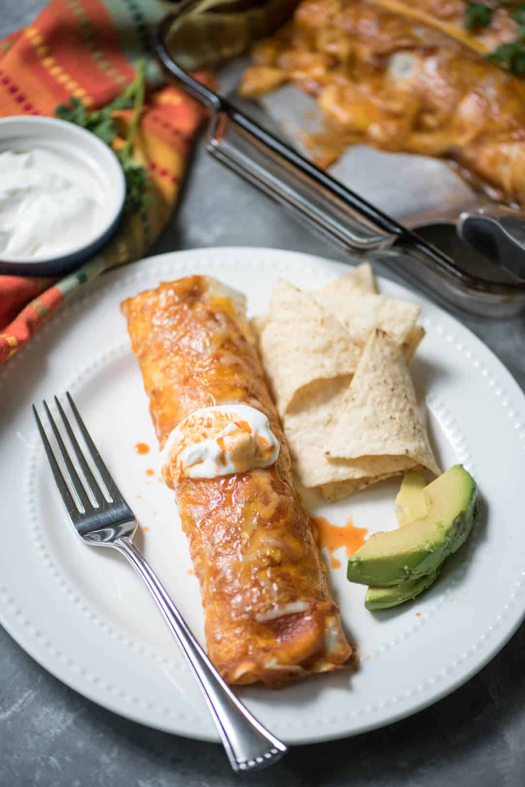 An enchilada topped with sour cream on a white plate with slices of avocado and tortilla chips.