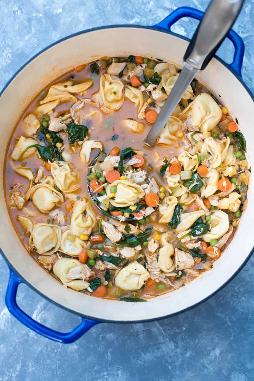A large blue Dutch oven filled with Chicken Vegetable Tortellini Soup and a ladle.