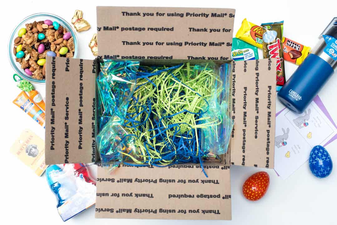 Easter grass and blue cellophane in a box with care package items around it.