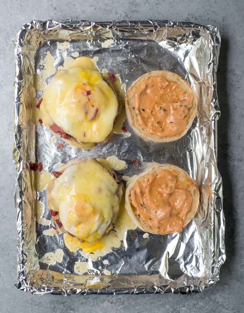 English muffin halves topped with sauce, corned beef, an egg, and cheese on a foil lined baking sheet.