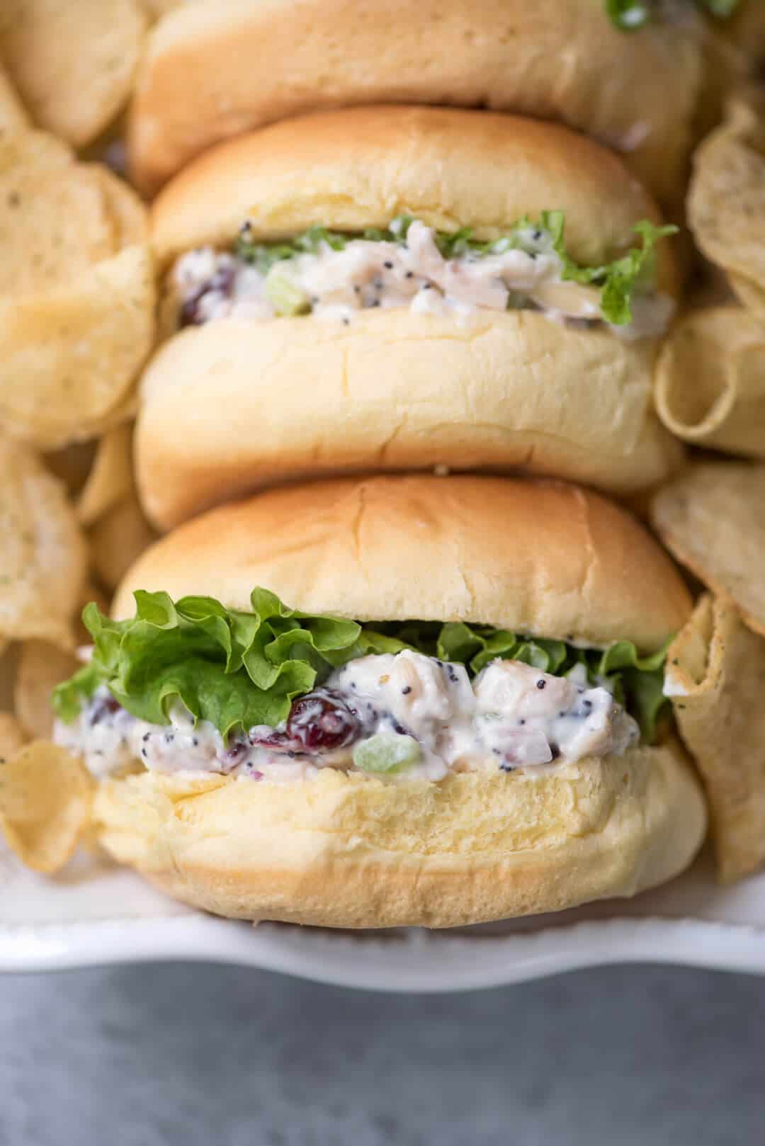 Cranberry Almond Poppy Seed Chicken Salad on potato buns with green leaf lettuce in a white serving dish with potato chips.