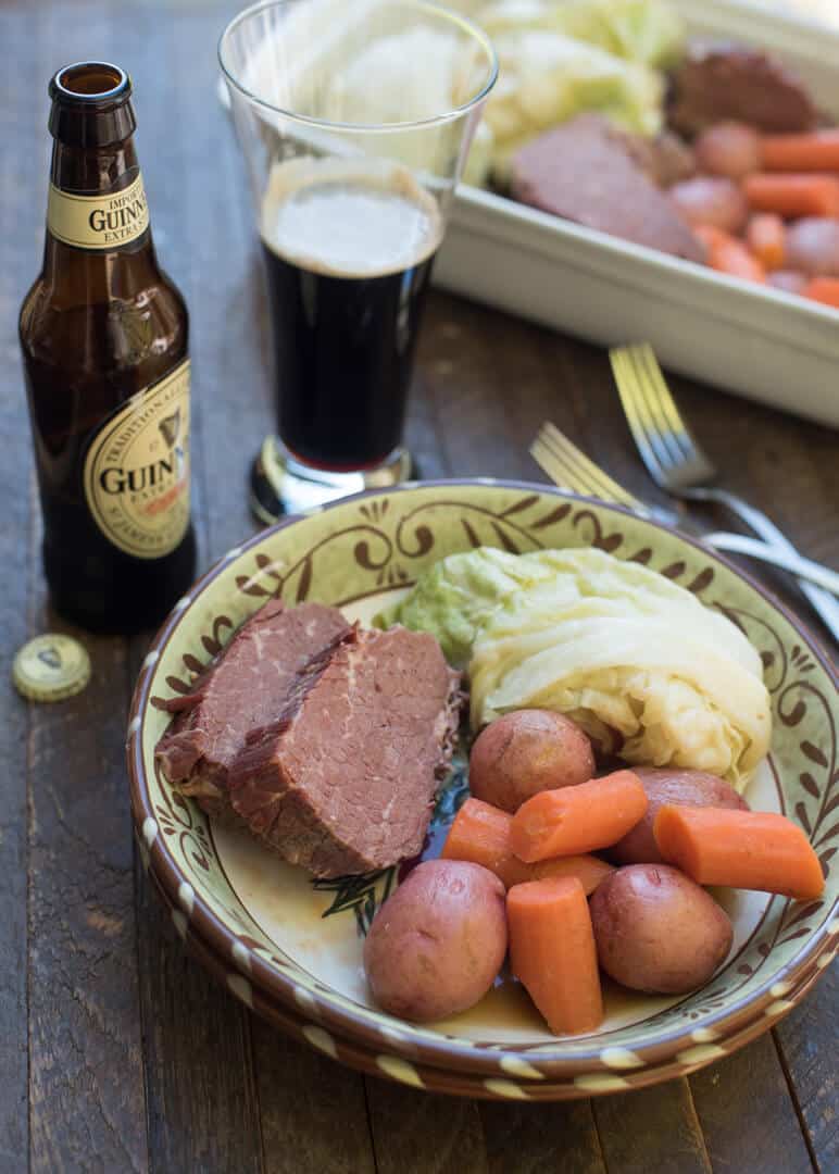 A bowl filled with slices of corned beef, potatoes, carrots, and cabbage with a bottle of Guinness in the background.