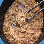 Metal tongs lifting shredded BBQ chicken from a slow cooker.