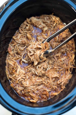 Metal tongs lifting shredded BBQ chicken from a slow cooker.