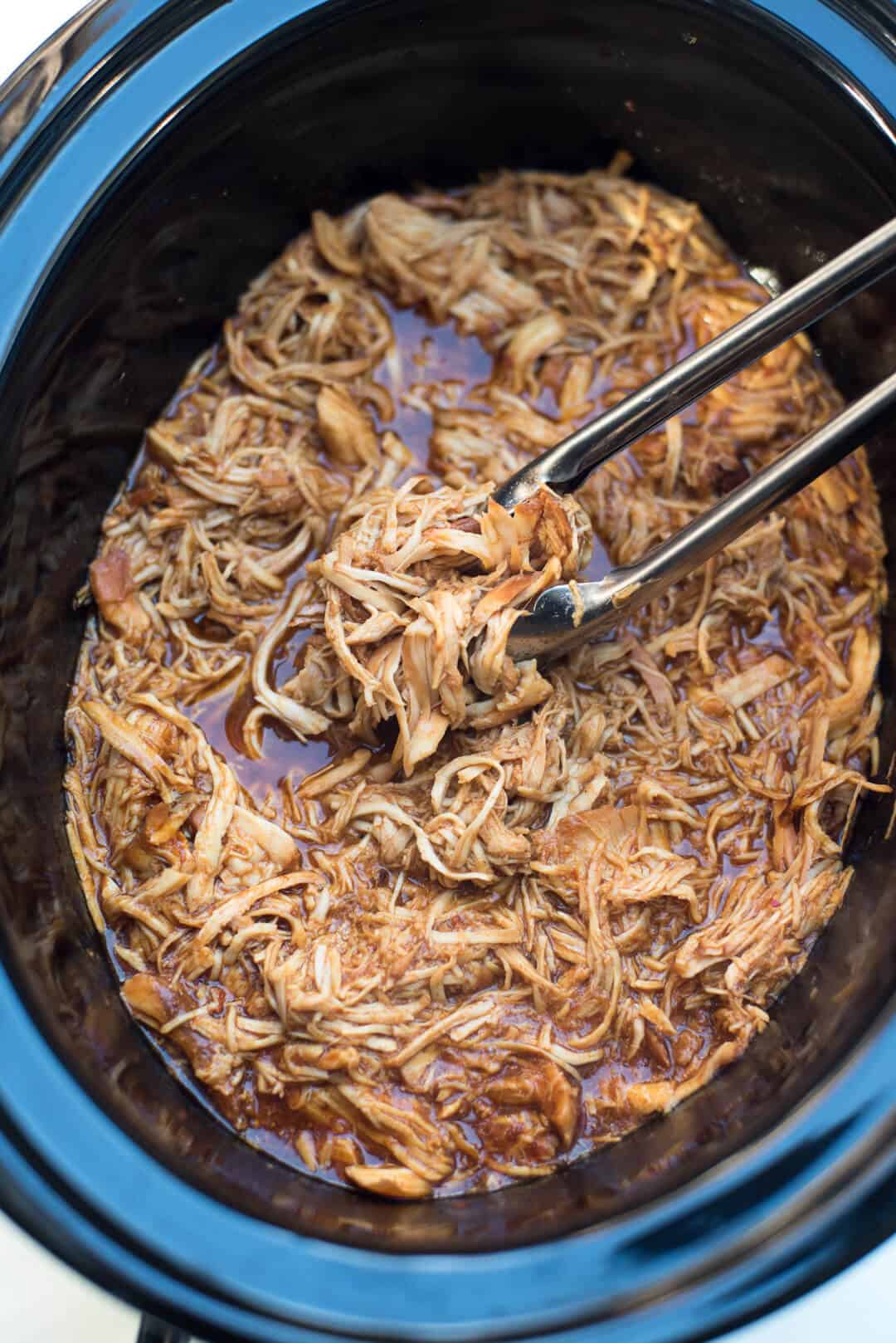 An over the top shot of the shredded BBQ Chicken in the slow cooker with tongs.