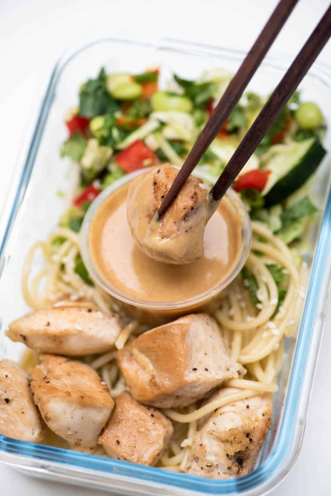 Chopsticks dip a piece of chicken into peanut sauce in the meal prep container.