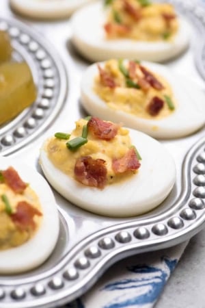 A close up of a deviled egg topped with crumbled bacon and chives on a silver platter.