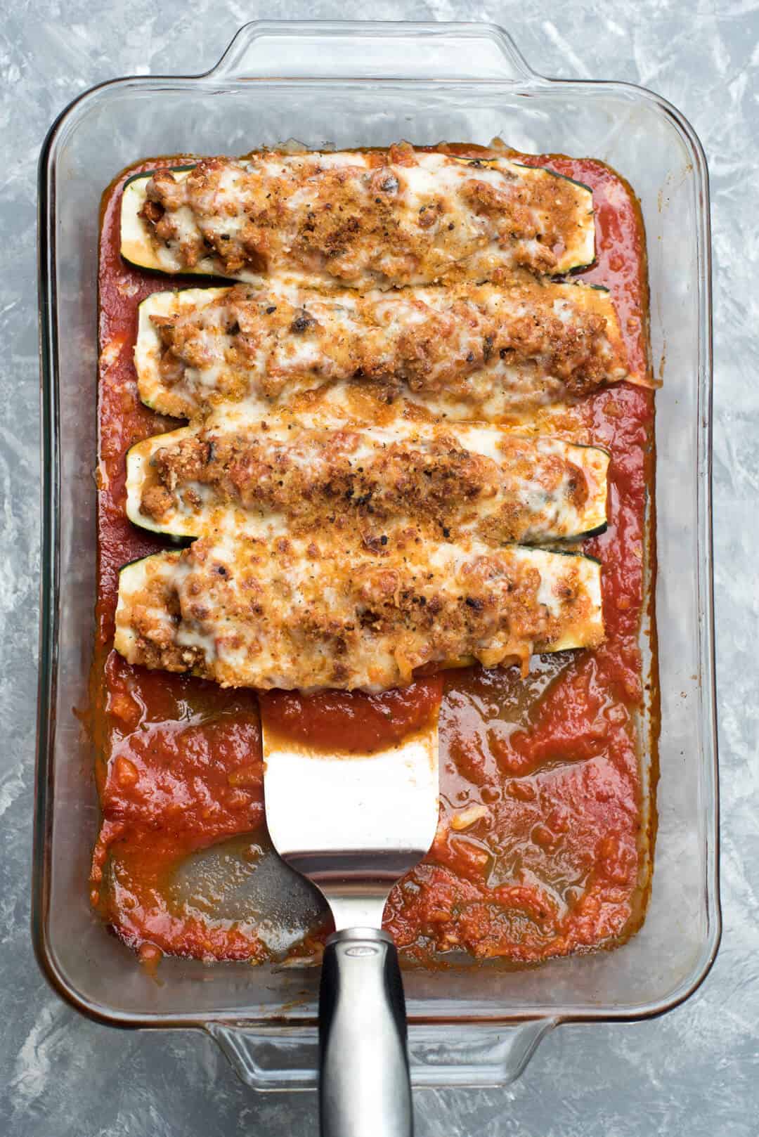 A spatula scoops up a zucchini boat from a casserole dish.