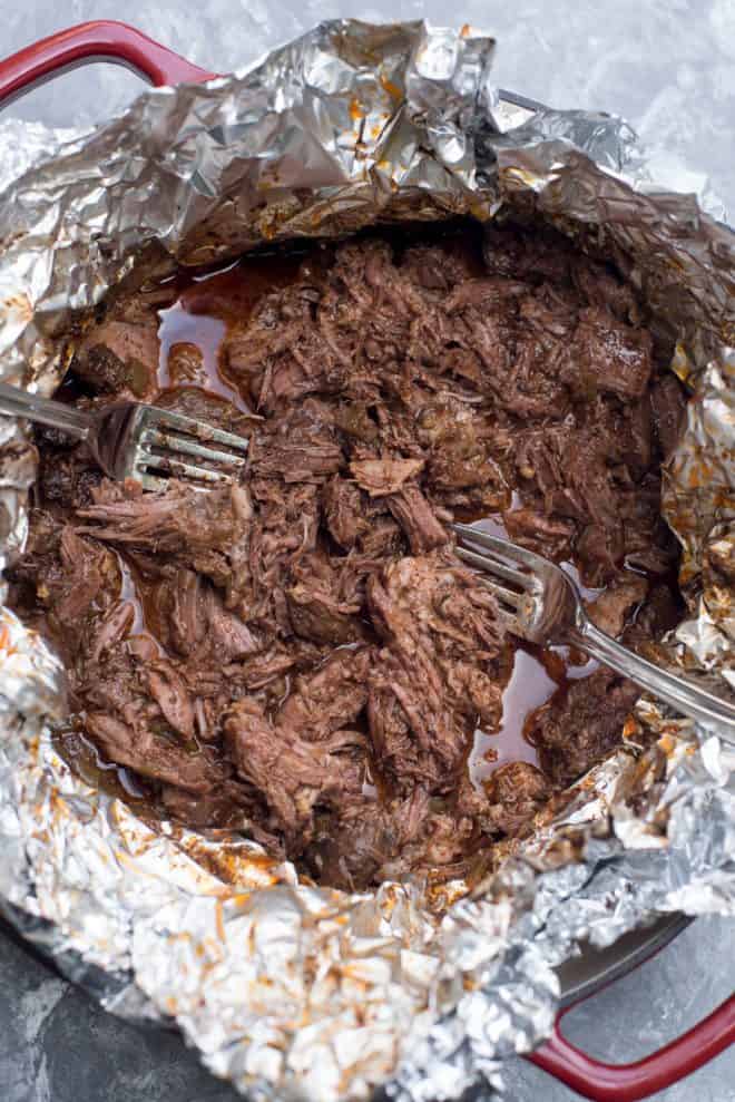 Two forks shredding Shredded Mexican Beef in a foil lined pan.