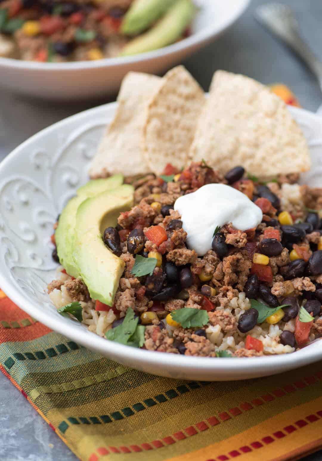 Brown rice topped with a cooked ground turkey mixture topped with sour cream and avocado in a white bowl.