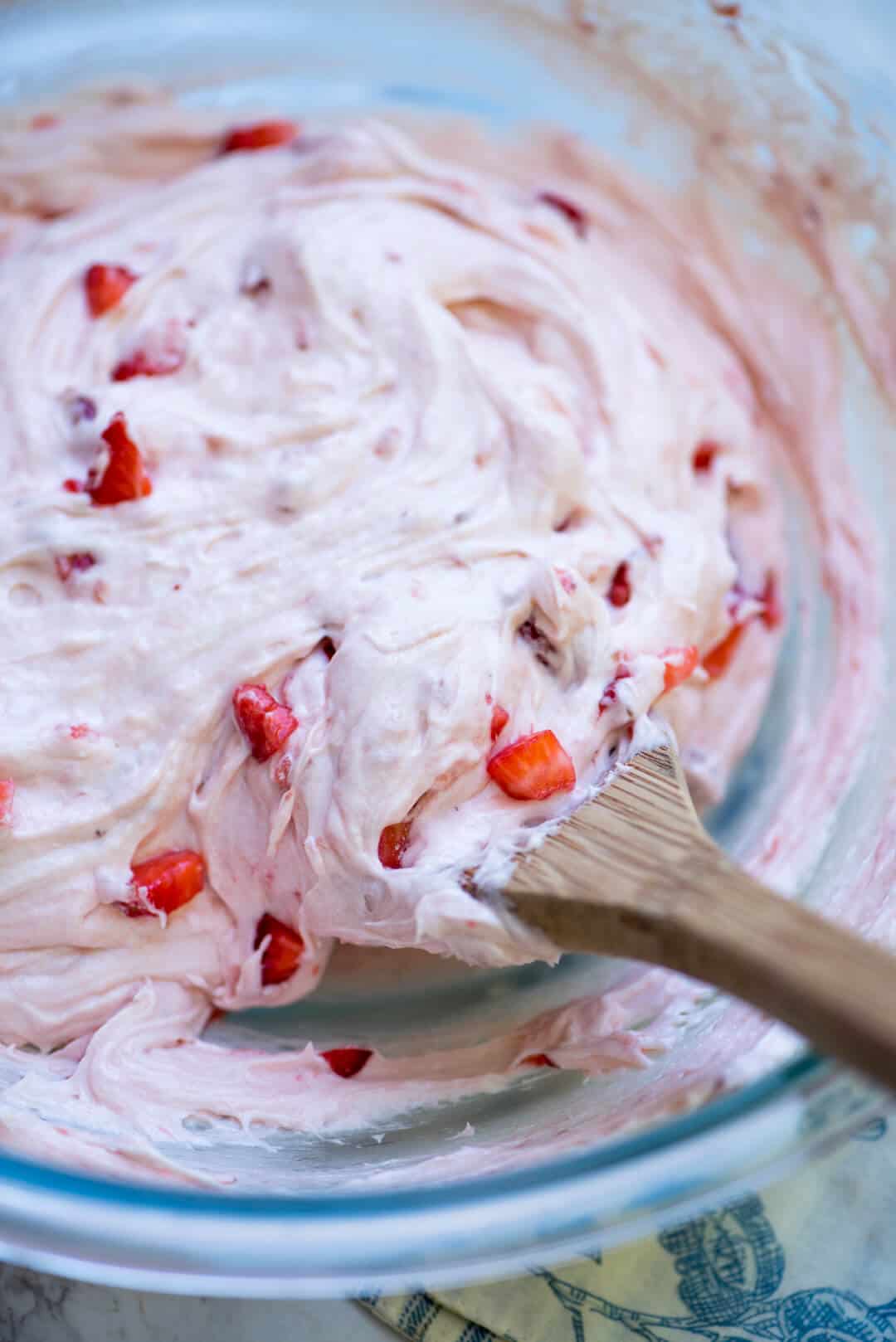 A wooden spoon stirs fresh strawberries into buttercream frosting.