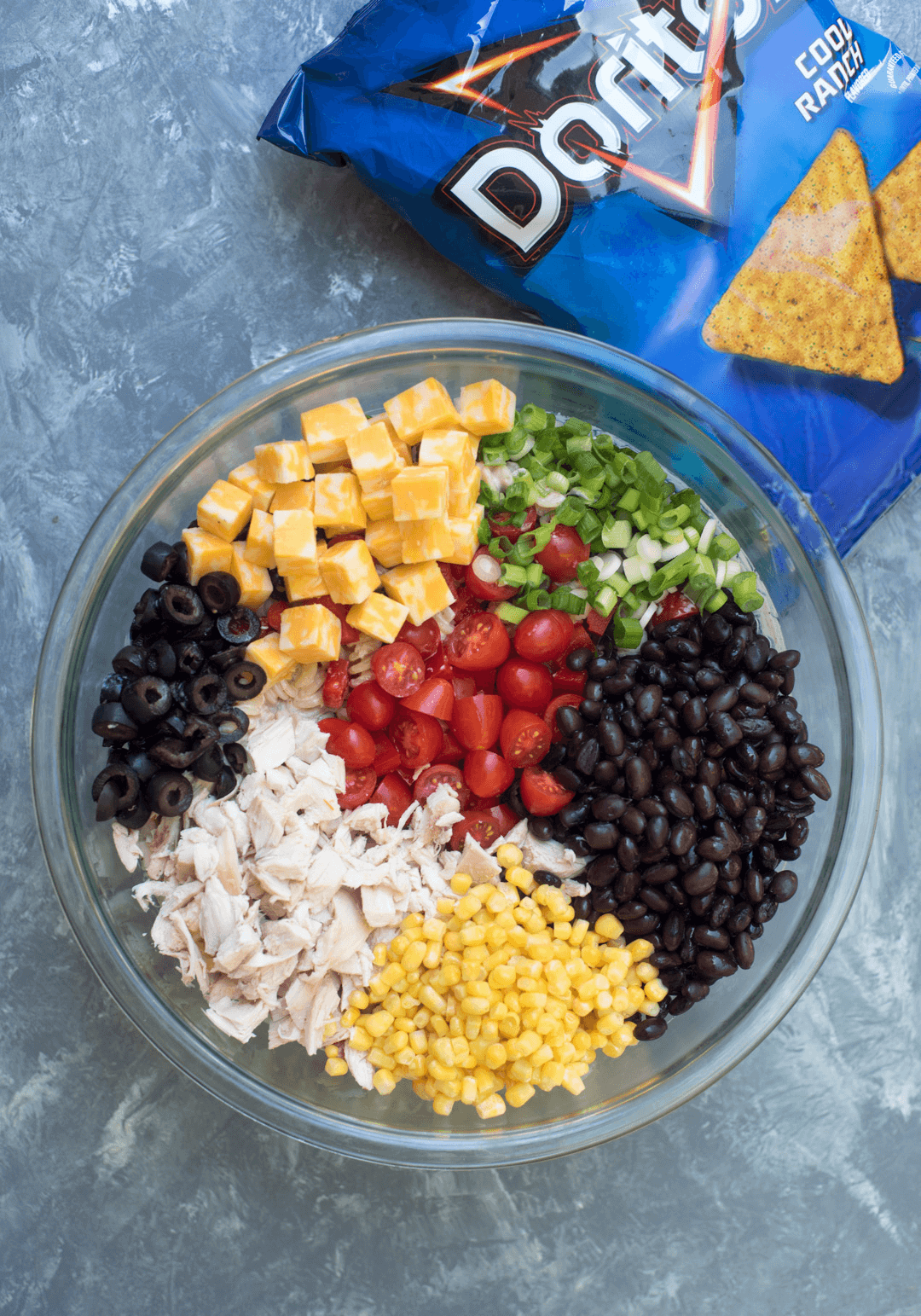 All of the ingredients for making the pasta salad set in rows in a mixing bowl.