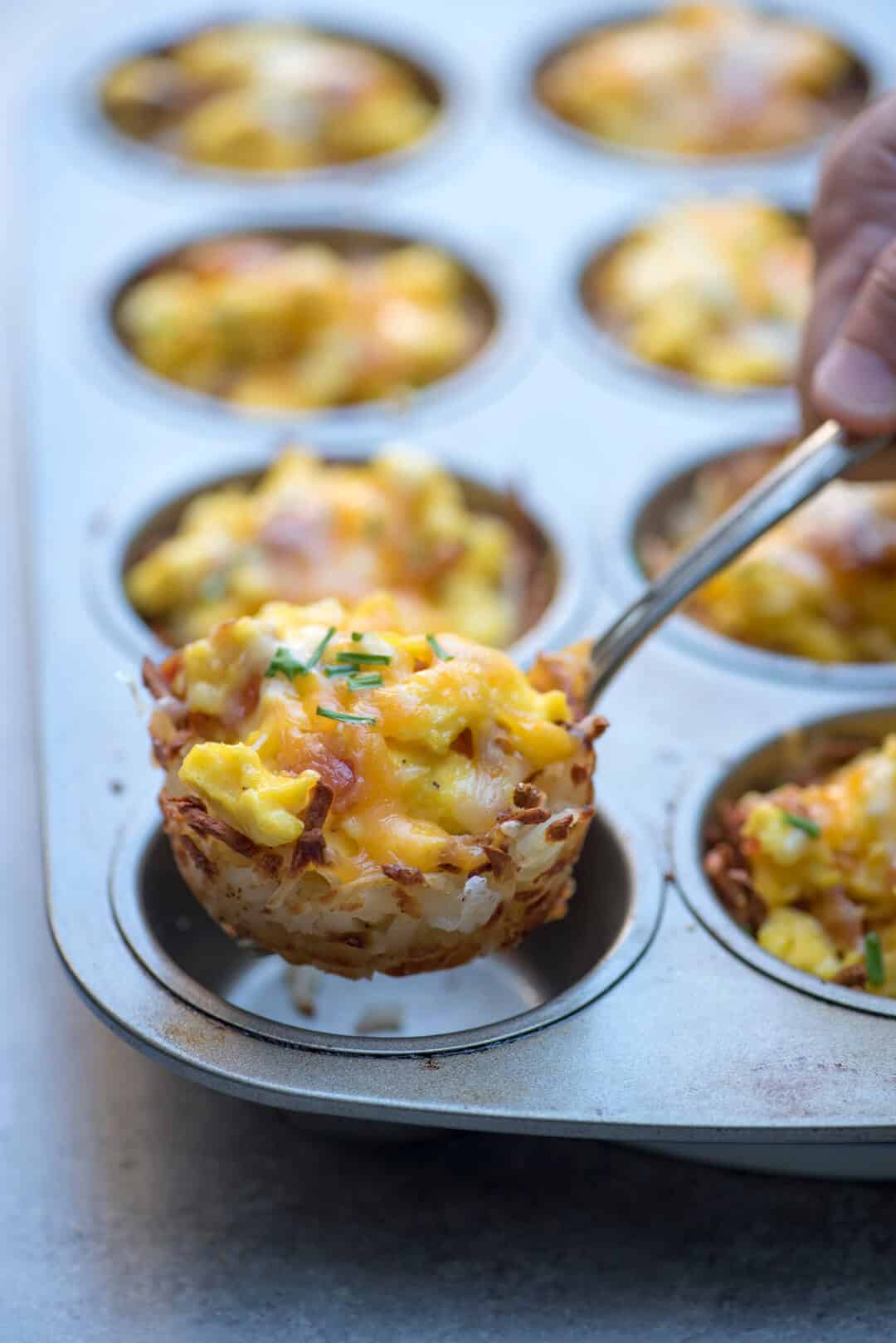 A fork lifts a Muffin Tin Hash Brown and Scrambled Egg Cup from a muffin pan.