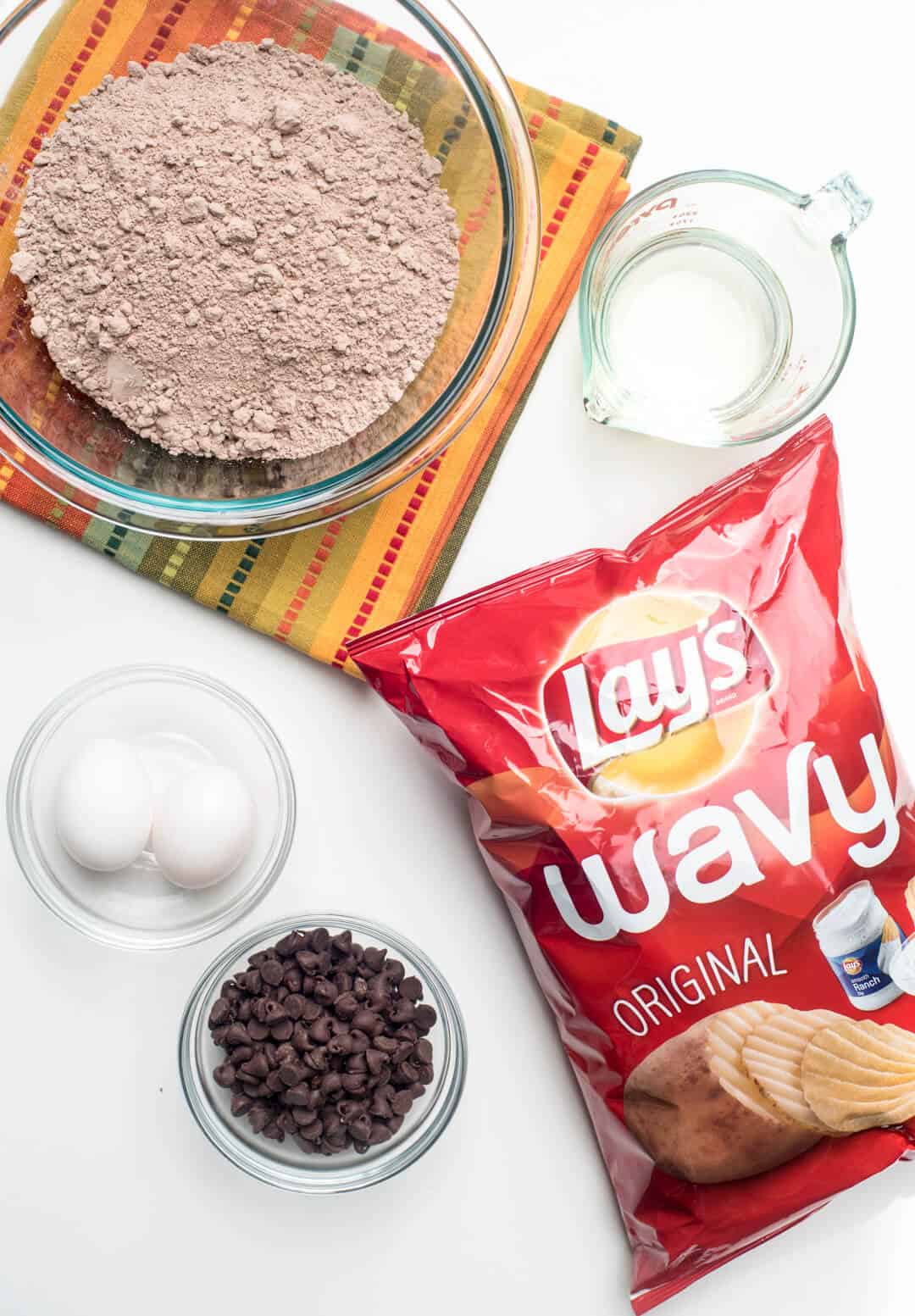 Boxed brownie mix in a bowl, a bag of Wavy Lay's Potato Chips and other ingredient on a white surface.