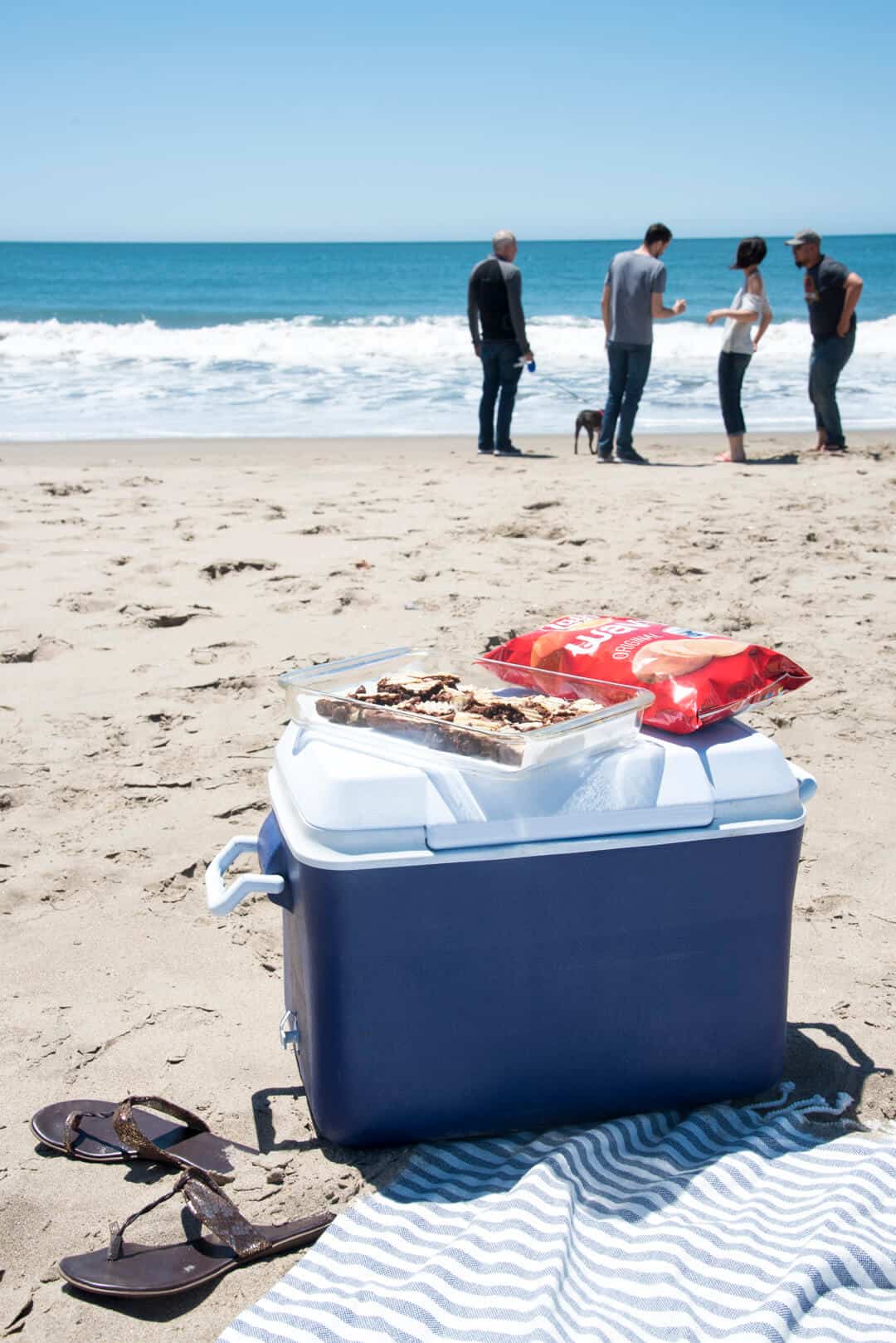 A cooler on the sand at the beach with a baking dish of brownies and potato chips on top.