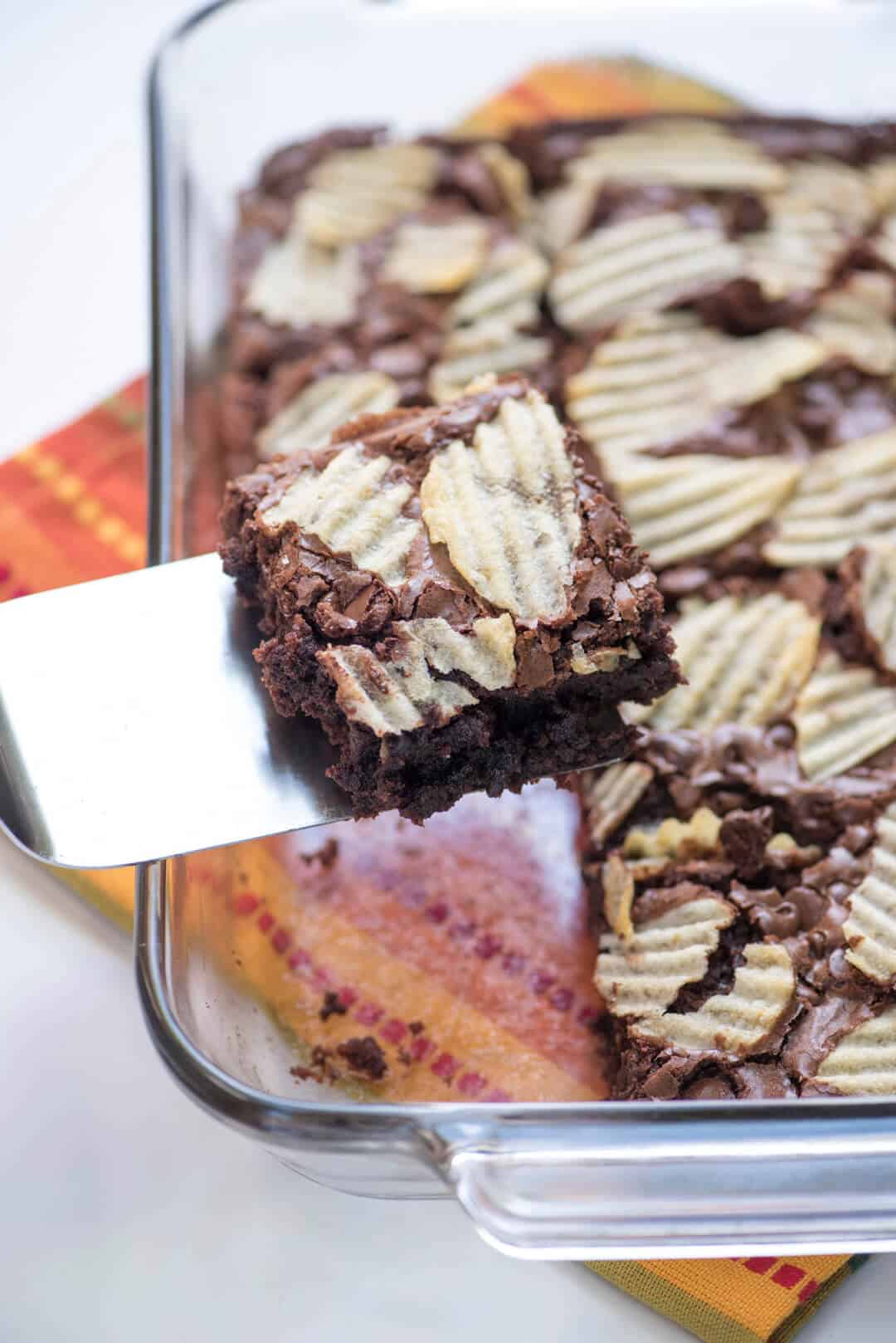 A spatula lifts a brownie topped with potato chips from a baking dish.