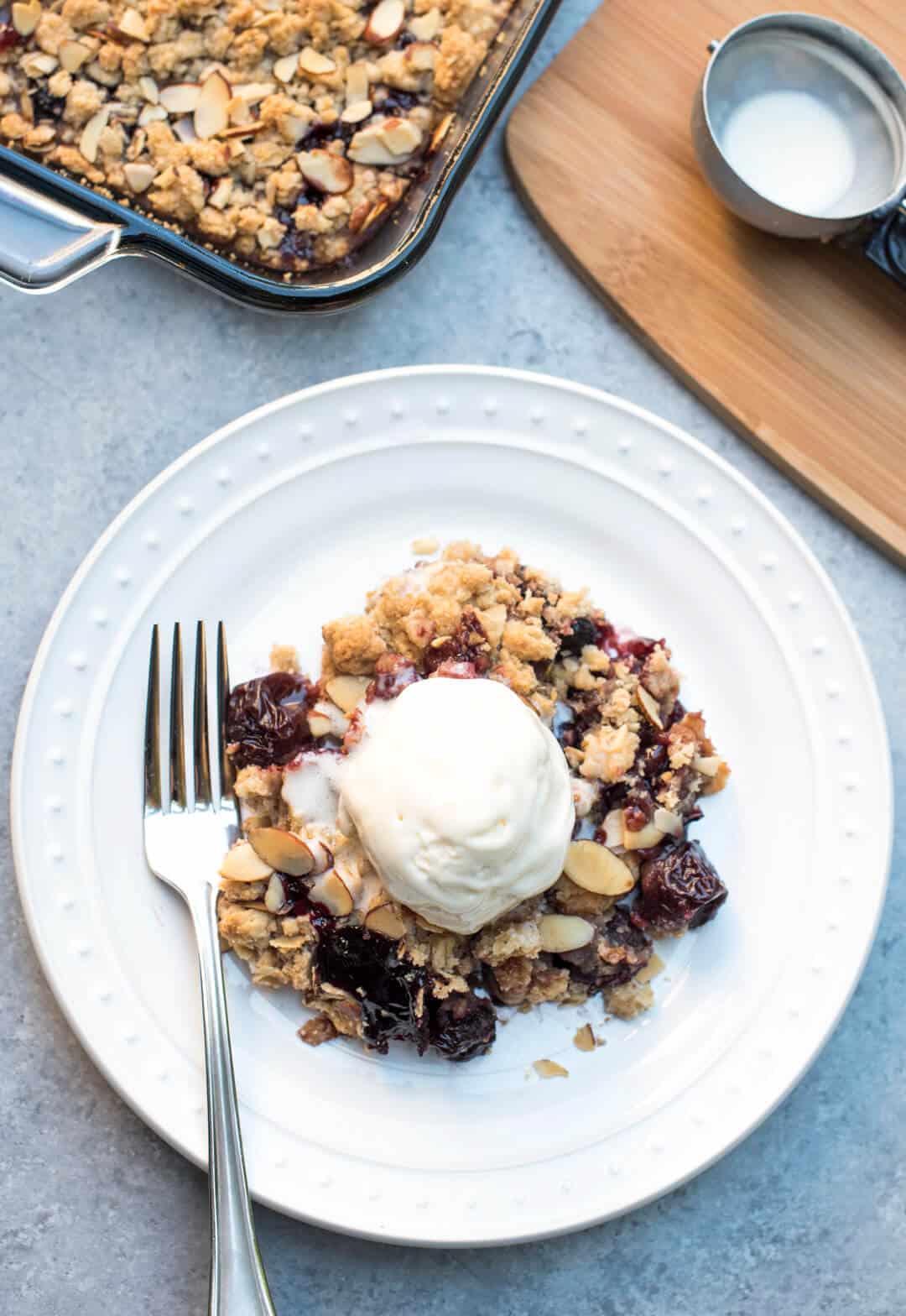 An over the top image of a serving of cherry crisp on a white plate with a scoop of vanilla ice cream and a fork.