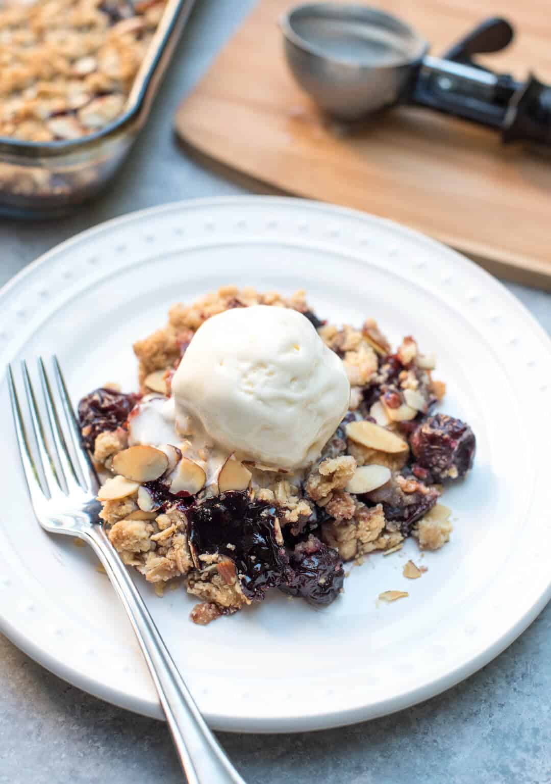 A serving of Cherry Crisp on a white plate with a scoop of ice cream and a fork.