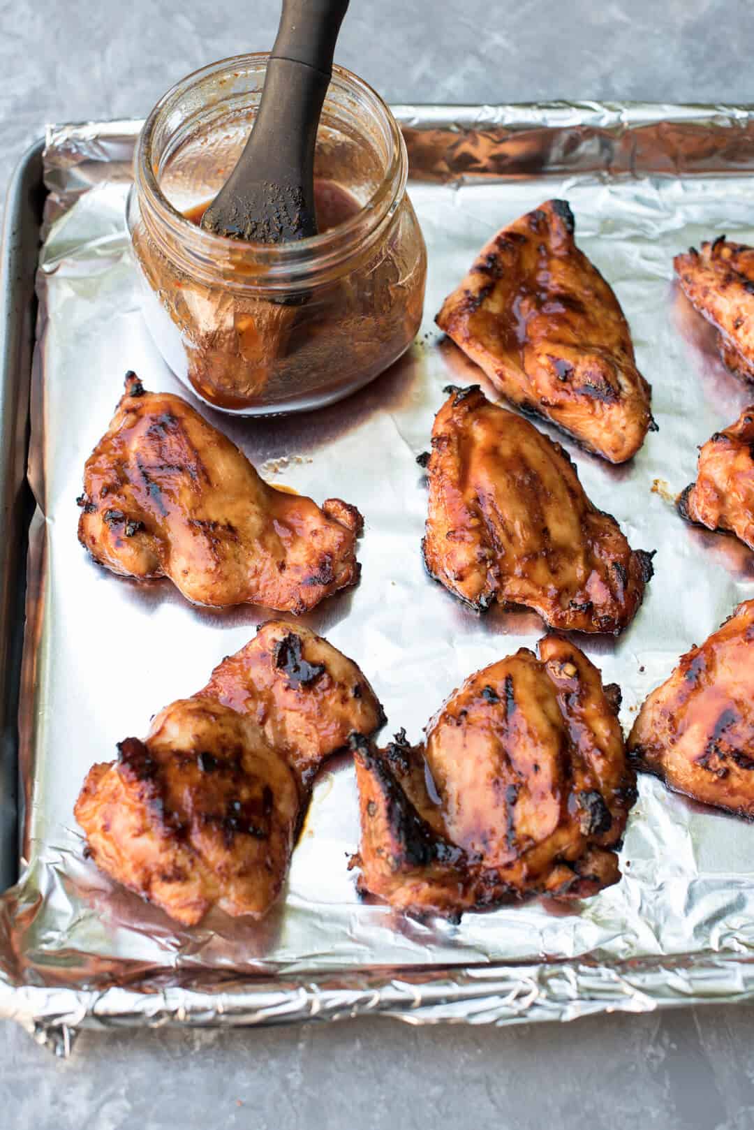 The grilled chicken on a foil lined baking sheet is basted with sauce.