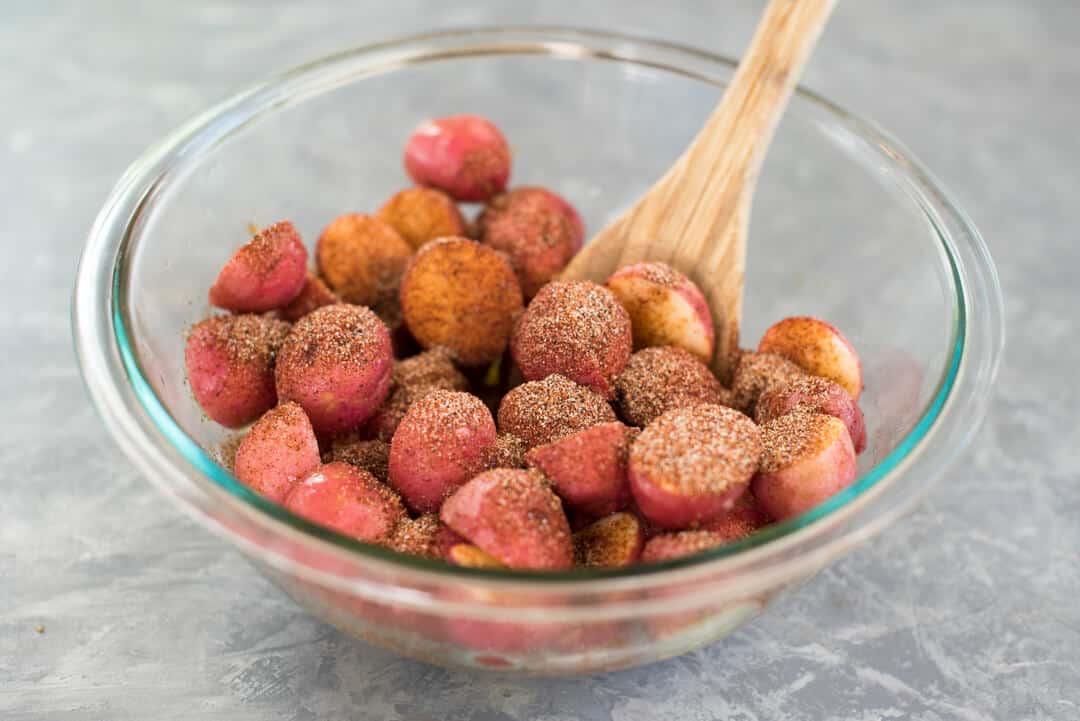 Halved mini red potatoes in a glass mixing bowl with coated olive oil and the BBQ spice mixture.