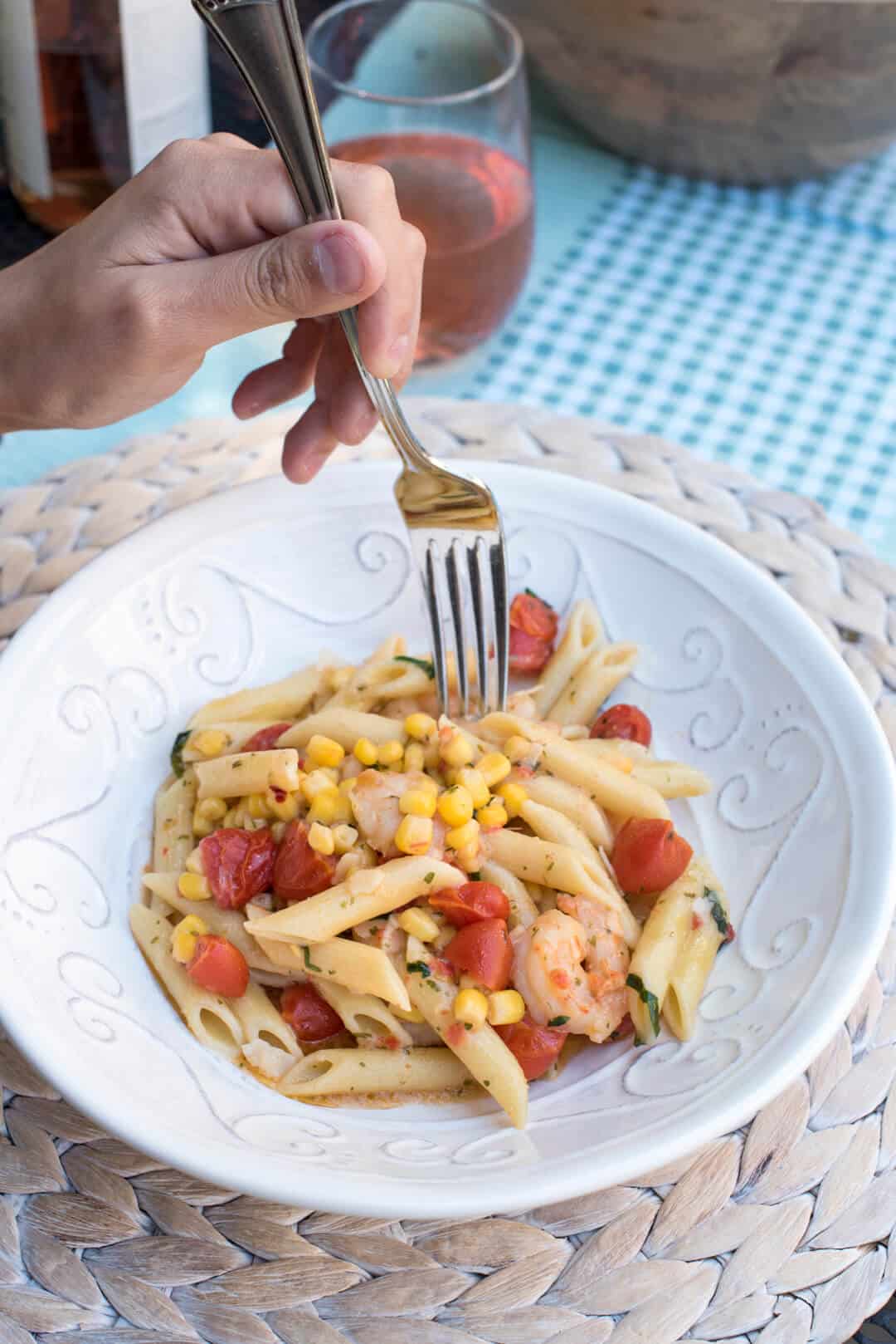 A fork presses into a bowl of pasta with shrimp, tomatoes, and corn.