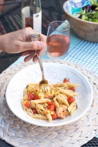 A hand pushes a fork into a bowl of penne with shrimp scampi and tomatoes.